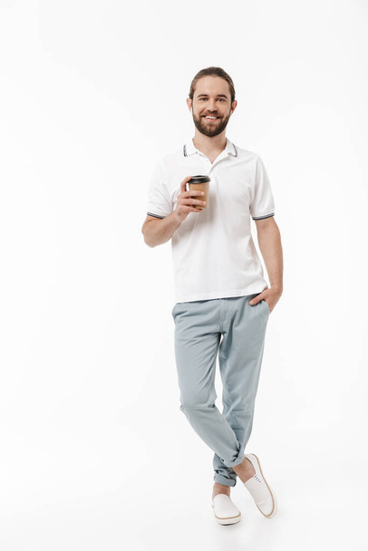 Photo of a young cheery handsome bearded man walking isolated over white wall background listening music with earphones drinking coffee. - Zdjęcie, obraz
