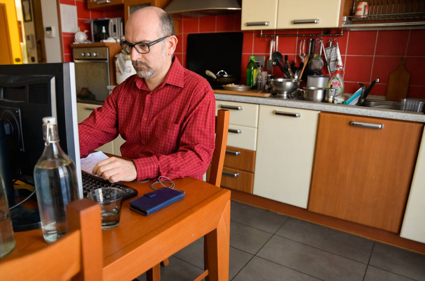 Turijn, Piemonte, Italië. april 2020. Coronavirus pandemie, een blanke man heeft een werkstation opgezet in de keuken. Op de keukentafel een monitor het toetsenbord en de smartphone met oortjes. - Foto, afbeelding