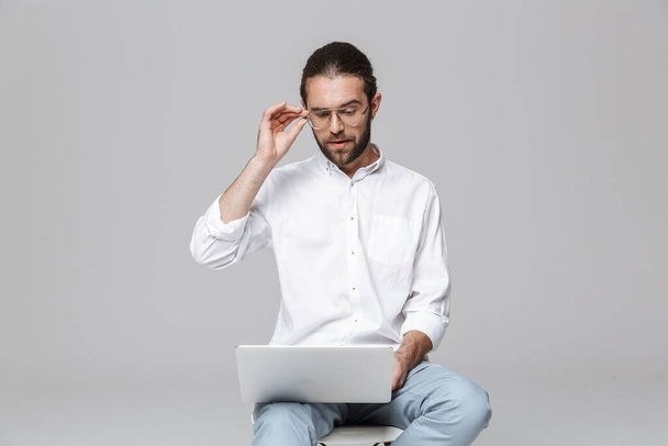 Image of a young concentrated serious handsome bearded man posing isolated over grey wall background wearing glasses using laptop computer. - 写真・画像