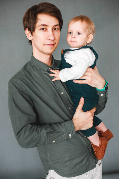 a one-year-old son sits in the arms of a young father on a gray background - 写真・画像