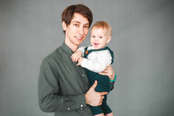 a one-year-old son sits in the arms of a young father on a gray background - Foto, Imagem