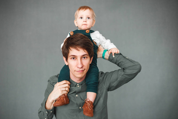 a one-year-old son sits on the shoulders of a young father on a gray background - Фото, изображение