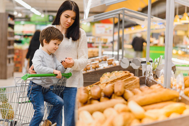 Cute child with mother choosing baked goods or pastry in bakery shop. Little boy with mom buying bread or buns at grocery supermarket - Photo, Image