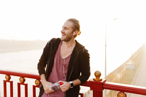 Handsome fit sportsman listening to music with wireless earphones on a bridge, holding water bottle - Fotoğraf, Görsel