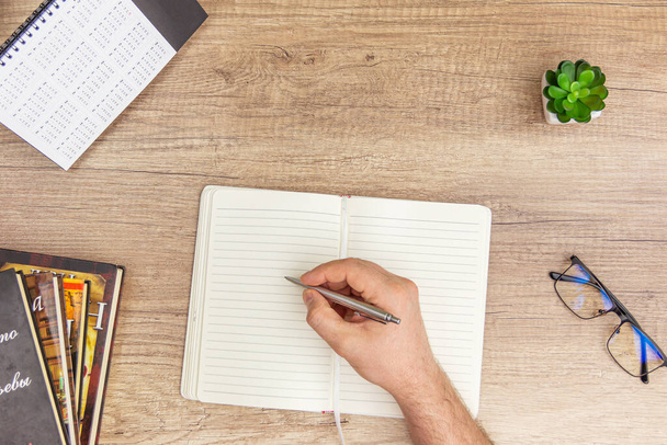 Αρσενικά χέρια γράφοντας σε ένα σημειωματάριο στο γραφείο με γυαλί, βιβλία, πυώδης σε ξύλινο φόντο - Φωτογραφία, εικόνα