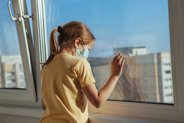 A little girl in a medical mask looks out the window. Coronavirus quarantine. - Φωτογραφία, εικόνα