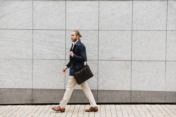 Attractive confident young bearded businessman walking outdoors in the street, carrying briefcase - Foto, imagen