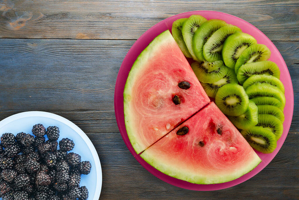watermelon and kiwi sliced on a red plate with fruits salad on a blue gray wooden background top view - Foto, immagini