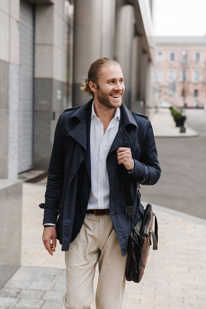 Attractive happy young bearded businessman walking outdoors in the street, carrying briefcase - Foto, immagini