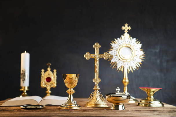 Catholic religion concept. Catholic symbols composition. The Cross, monstrance,  Holy Bible and golden chalice on wooden altar and gray background.  - Photo, Image