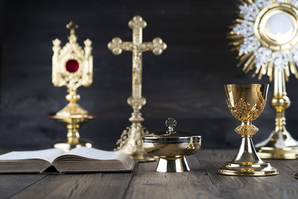 Katholieke religie concept. Katholieke symbolen samenstelling. Kruis, monstrans, bijbel en gouden kelk op houten altaar en grijze achtergrond.  - Foto, afbeelding