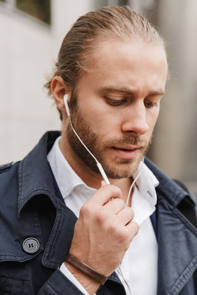 Image of handsome young confident businessman wearing earphones walking on city street - Foto, Bild