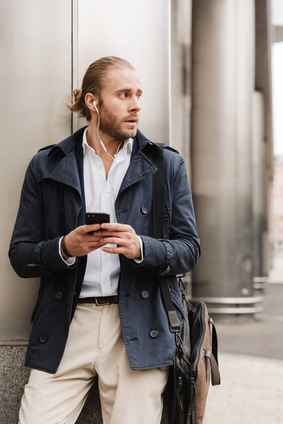 Attractive young blonde haired man in formal wear talking through earphones, holding mobile phone while standing outdoors on a city street - Foto, afbeelding