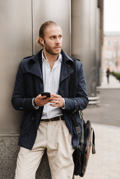Attractive young blonde haired man in formal wear talking through earphones, holding mobile phone while standing outdoors on a city street - Photo, Image