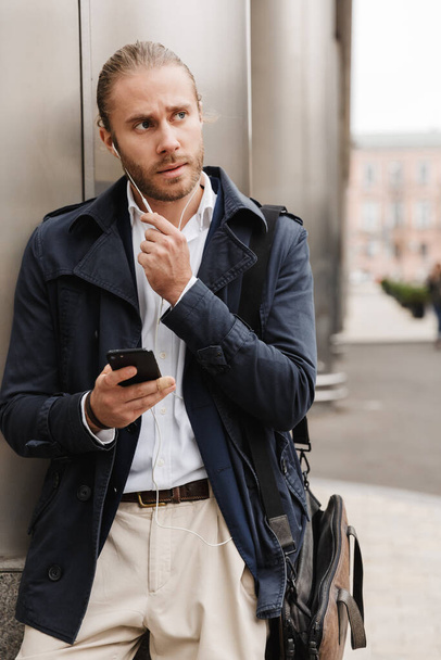 Attractive young blonde haired man in formal wear talking through earphones, holding mobile phone while standing outdoors on a city street - Foto, imagen