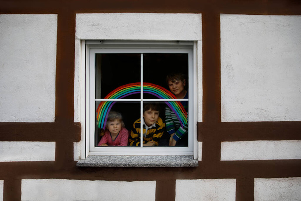 Three kids, two school kids boys and toddler girl with rainbow painted with colorful window color during pandemic coronavirus quarantine. Children painting rainbows with the words Lets all be well. - Photo, Image