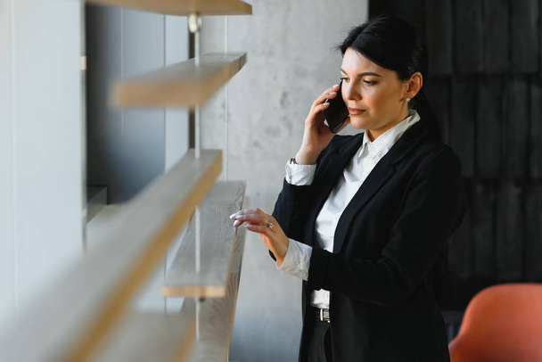 Vrolijke jonge zakenvrouw die in een modern kantoor staat te praten op haar mobiele telefoon. Mooi vrouwelijk model met behulp van mobiele telefoon. - Foto, afbeelding