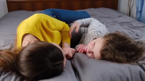 Mała dziewczynka przytula się całując szczęśliwą młodą mamusię leżącą w przytulnym łóżku, uśmiechniętą matkę lub nianię odpocząć wziąć, córka leżąca w łóżku cieszyć się chwilą czułości z mamą, miłość. Koncepcja rodziny krewnych - Materiał filmowy, wideo