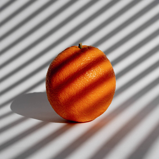 Ωρίμασε ζουμερό πορτοκάλι σε λευκό φόντο το πρωί με φως μέσα από τα λουτρά. Στρέμματα ηλιακού φωτός από τις περσίδες του παραθύρου. Ελάχιστος δημιουργικός σχεδιασμός - Φωτογραφία, εικόνα