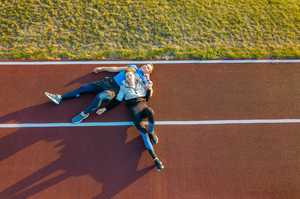 Widok z góry na dół z lotu ptaka dwóch młodych sportowców i sportowców leżących na czerwonej gumie biegających po boisku stadionowym odpoczywających latem po maratonie joggingowym. - Zdjęcie, obraz