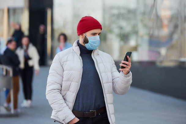 A man with a beard wearing a medical face mask to avoid the spread coronavirus (COVID-19). A guy with a surgical mask on the face using a smartphone in the center of the city. - 写真・画像