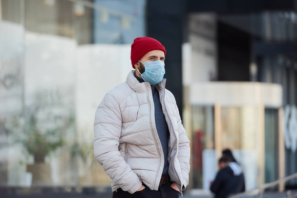 Um homem caucasiano usando uma máscara facial médica para evitar a propagação do coronavírus (COVID-19). Um tipo com uma máscara cirúrgica na cara que fica numa rua.
. - Foto, Imagem