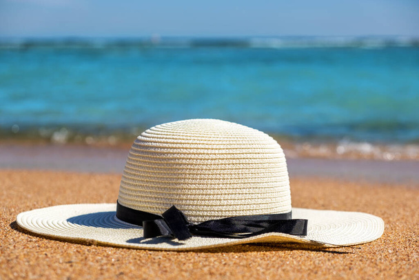 Chapeau de paille femme blanche posée sur la plage de sable tropical avec de l'eau bleue vibrante de l'océan en arrière-plan par une journée d'été ensoleillée. Concept de voyage vacances et destination. - Photo, image