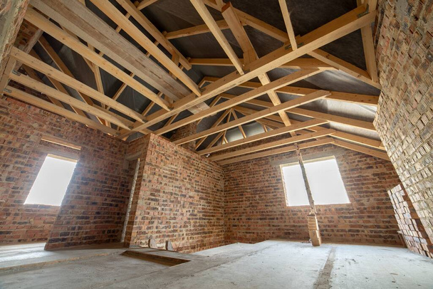 Wnętrze niedokończonego murowanego domu z betonową podłogą, gołe ściany gotowe do tynkowania i drewniany poddasze ramy dachowej w budowie. - Zdjęcie, obraz