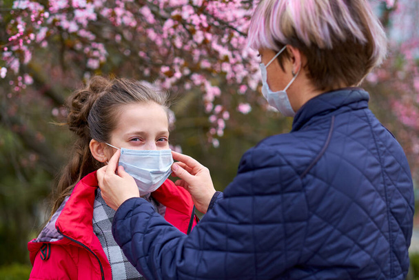 Mutter und Tochter mit Gesichtsmaske sind in der Stadt im Freien, blühende Bäume, Frühlingszeit, Blütezeit - Konzept für Allergien und Gesundheitsschutz vor staubiger Luft - Foto, Bild