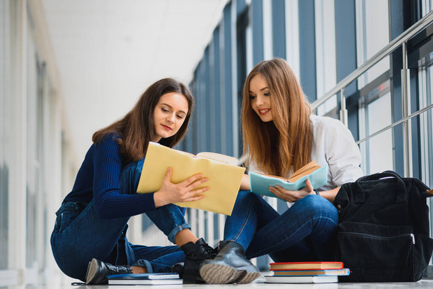 two pretty female students with books sitting on the floor in the university hallway - Фото, изображение
