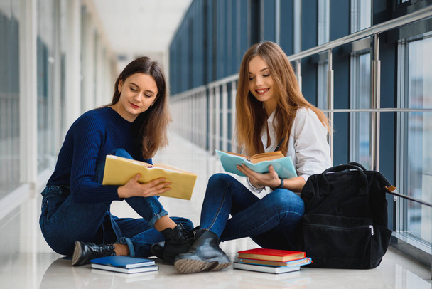 two pretty female students with books sitting on the floor in the university hallway - Фото, зображення