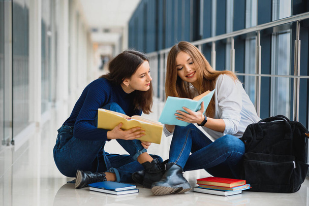 two pretty female students with books sitting on the floor in the university hallway - Фото, изображение