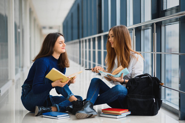 two pretty female students with books sitting on the floor in the university hallway - Zdjęcie, obraz