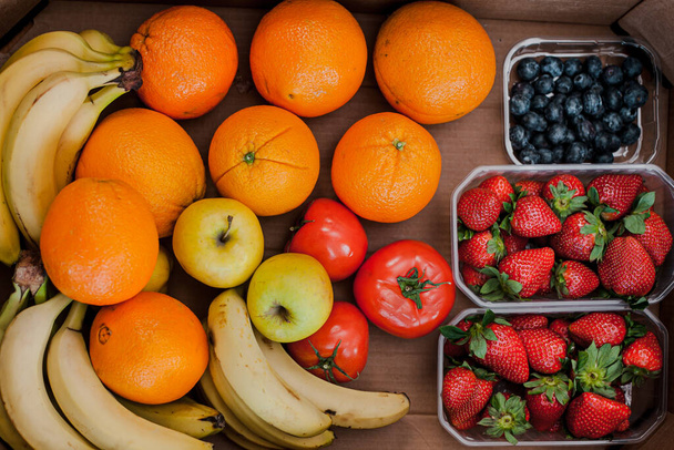 Άποψη της παράδοσης φρέσκων βιολογικών λαχανικών σε κουτί, τοπική αγορά τροφίμων. Φρέσκα και πολύχρωμα λαχανικά και φρούτα σε ένα χαρτόνι. Πορτοκάλια, μπανάνες, μήλα, ντομάτες, φράουλες και μύρτιλλα. - Φωτογραφία, εικόνα