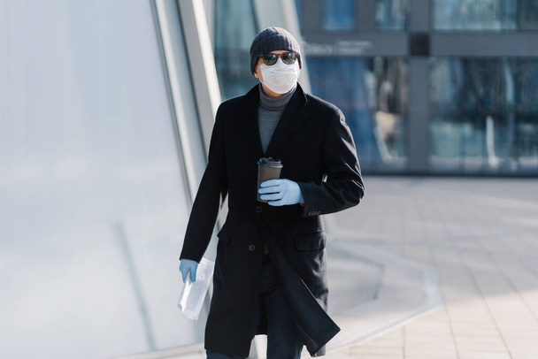 Outdoor shot van de jongeman in zwarte jas, steriel medisch masker en beschermende rubberen handschoenen, drinkt afhaalkoffie, heeft papieren, moet gaan werken poses op lege straat tijdens quarantaine, coronavirus - Foto, afbeelding