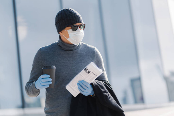 Outdoor image of male worker returns home from work, turns head aside, drinks coffee to go, holds newspaper, wears medical sterile mask and rubber gloves, prevents coronavirus spread. Infectious virus - Photo, Image