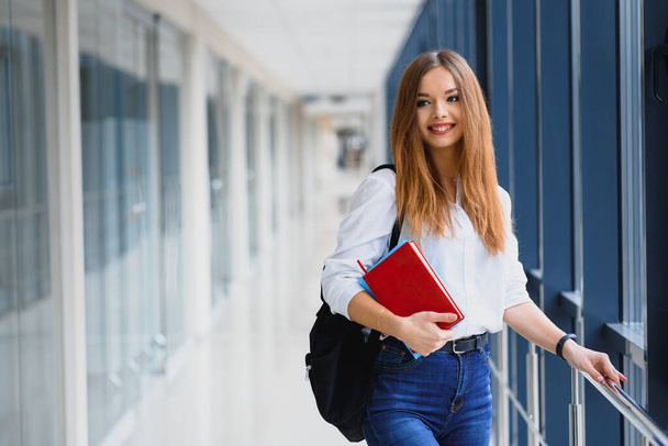 Positivity beautiful girl smiling at camera, standing on corridor with notes as backpack, going to lesson. Happy brunette female student studying in luxury university. - Fotó, kép