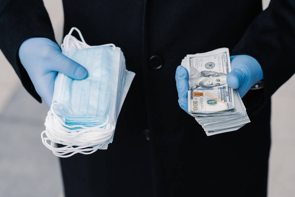 Increasing prices on medical masks durig spread of coronavirus epidemic. Person in protective medical gloves holds stack of masks and much money. Profitable business, speculation during quarantine. - Photo, Image