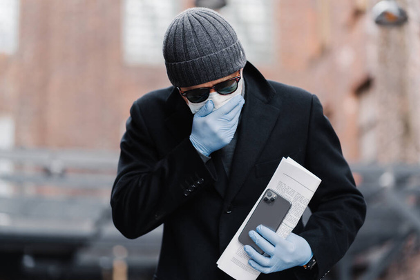 Infected young man has sneezing and coughing, wears disposable protective mask against covid-19, holds modern smartphone and papers, poses outdoor against blurred background. Global pandemic concept - Фото, изображение