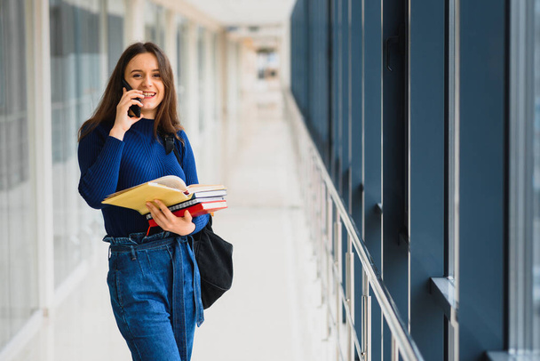 Cheerful brunette student girl with black backpack holds books in modern building. female student standing with books in college hallway - Foto, imagen