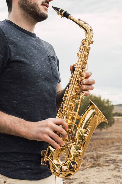 Young bearded man plays saxophone in a natural place during a cloudy day in the countryside. Playing musical instrument in the nature. - Foto, Imagen
