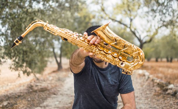 Junger Mann mit einem Saxophon vor dem Gesicht auf einem Felsweg an einem natürlichen Ort. Saxofonist mit Saxofon, Unschärfeeffekt im Hintergrund. - Foto, Bild