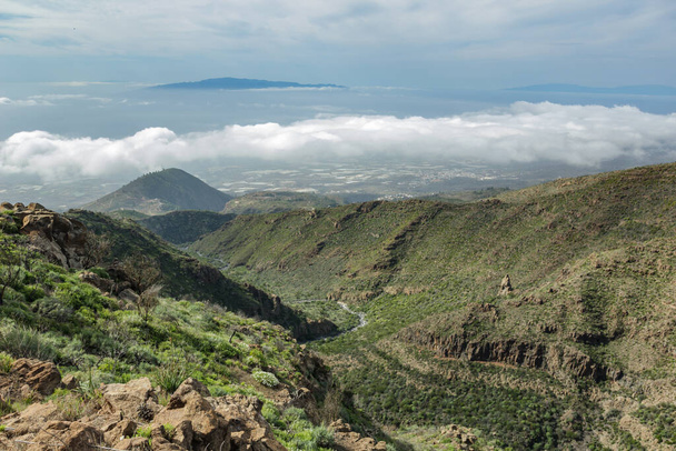 Tenerife 'nin batı yakasının panoramik manzarası ve bulutların üzerindeki La Gomera adası. Dağ yolunda yürüyüş. Etrafı lava kayaları ve bitki örtüsüyle çevrili.. - Fotoğraf, Görsel