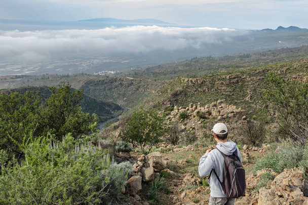 Ένας νεαρός άνδρας με ένα καπέλο με ένα σακίδιο πίσω από την πλάτη του ταξιδεύει κατά μήκος μιας διαδρομής στο Εθνικό Πάρκο Teide. Πεζοπορία από το ορεινό μονοπάτι που περιβάλλεται από ενδημική βλάστηση και χωράφια με πέτρες λάβας. Τενερίφη. - Φωτογραφία, εικόνα