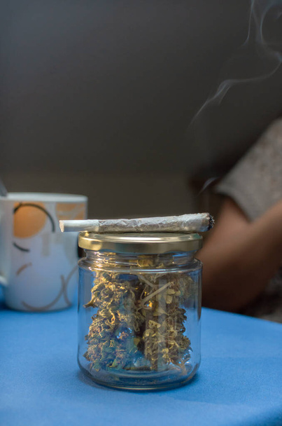 People smoking a marijuana joint whit a cup of coffee at home. Reuse glass jar full of cannabis buds with a weed cigarette, close up. Copy space top. - Photo, Image