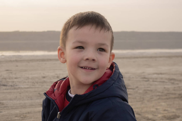 Chłopiec 4 lata w niebieskiej kurtce na pustej plaży. Dziecko się uśmiecha.. - Zdjęcie, obraz
