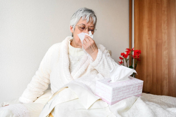 Coronavirus pandemia. Terveydenhuolto, flunssa, hygienia ja ihmisten käsite. Sairas vanhempi nainen paperipyyhkeellä puhaltaa nenäänsä kotona
. - Valokuva, kuva