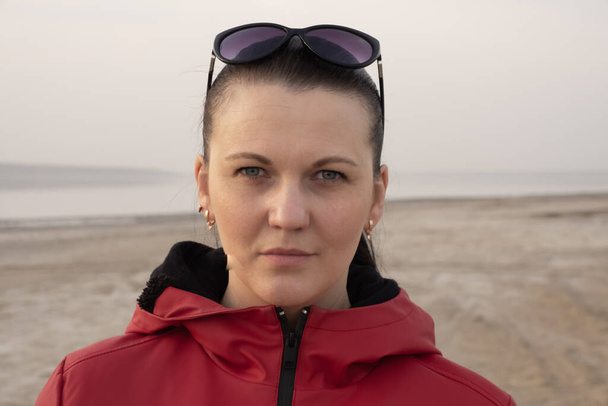 サングラスのブルネットの女性と隔離中の空のビーチの赤いジャケット. - 写真・画像