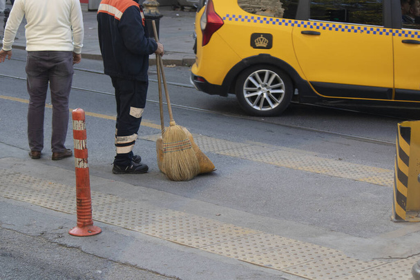 道路脇のアスファルトの上で清掃を行う市の労働者。彼の手にはほうきとシャベルがある. - 写真・画像