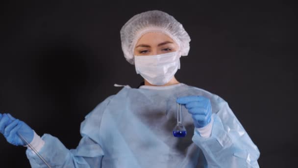A drop of blue liquid falls into a transparent vessel from a glass tube of a heat-resistant water-measuring water-indicating device. Young woman in protective clothing. On the face of a protective - Materiał filmowy, wideo